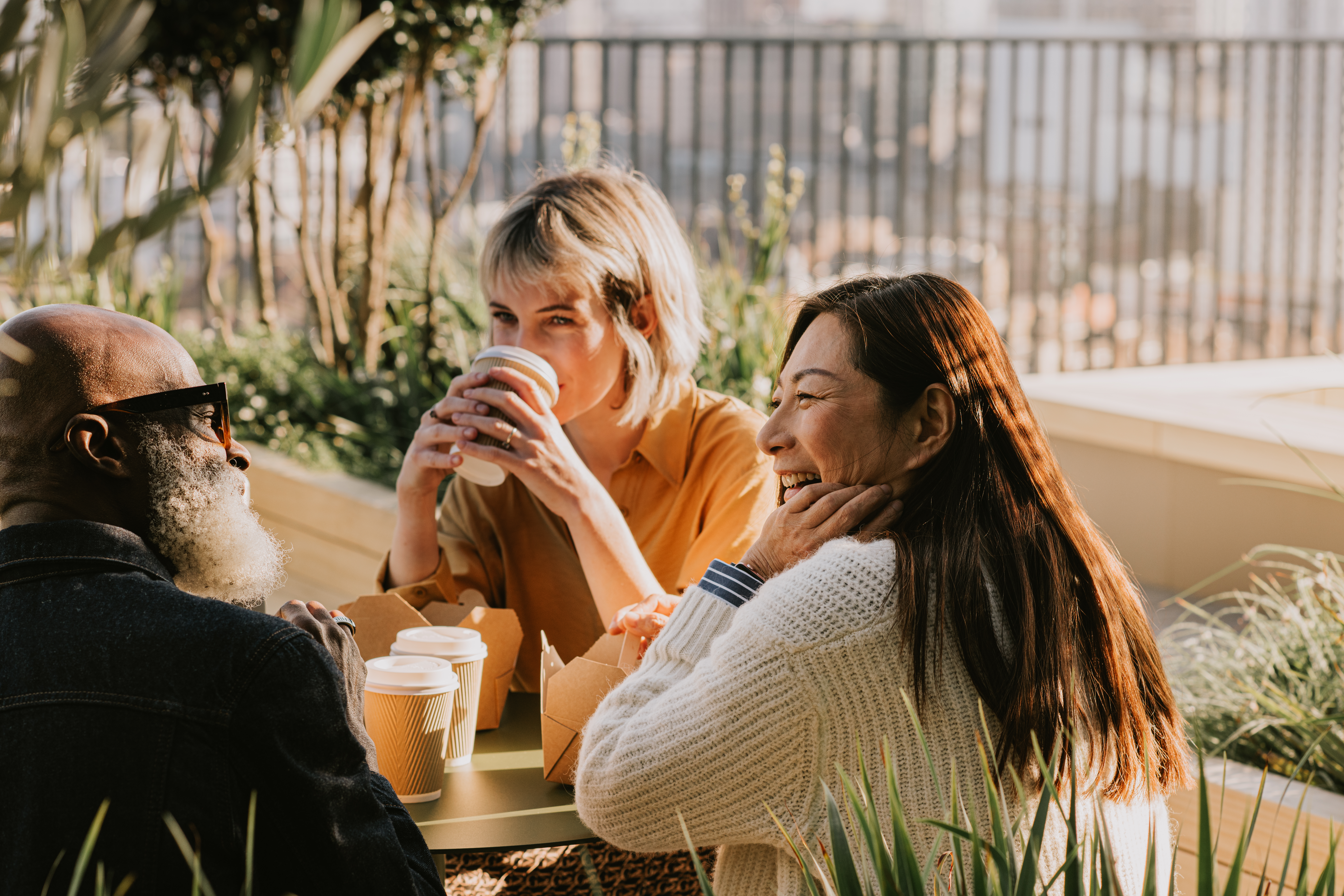 Three colleagues sit outside on a sunny day and enjoy a takeaway lunch and some hot drinks. They are comfortable with each other, having a light hearted discussion.