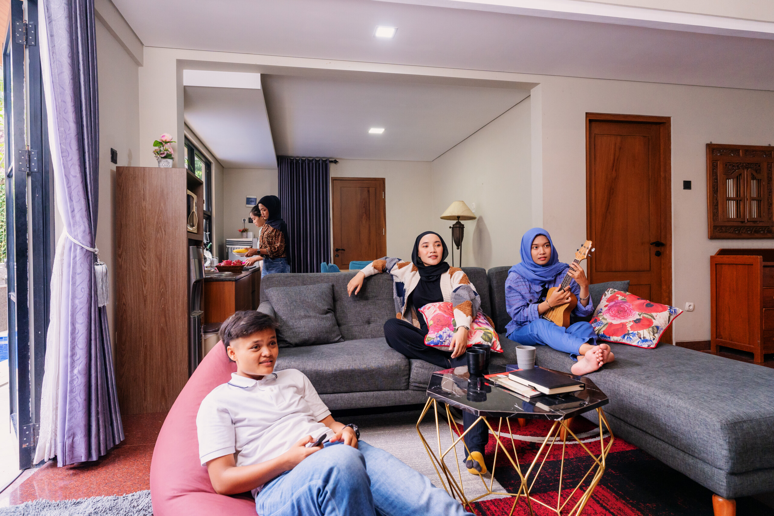 A group of young Muslim friends spending leisure time together in a stylish modern living room. One person is playing a ukulele, another is watching TV while lounging on a bean bag, and another is preparing food in the kitchen. The atmosphere is relaxed and cozy, with a mix of activities such as music, entertainment, and casual conversation.