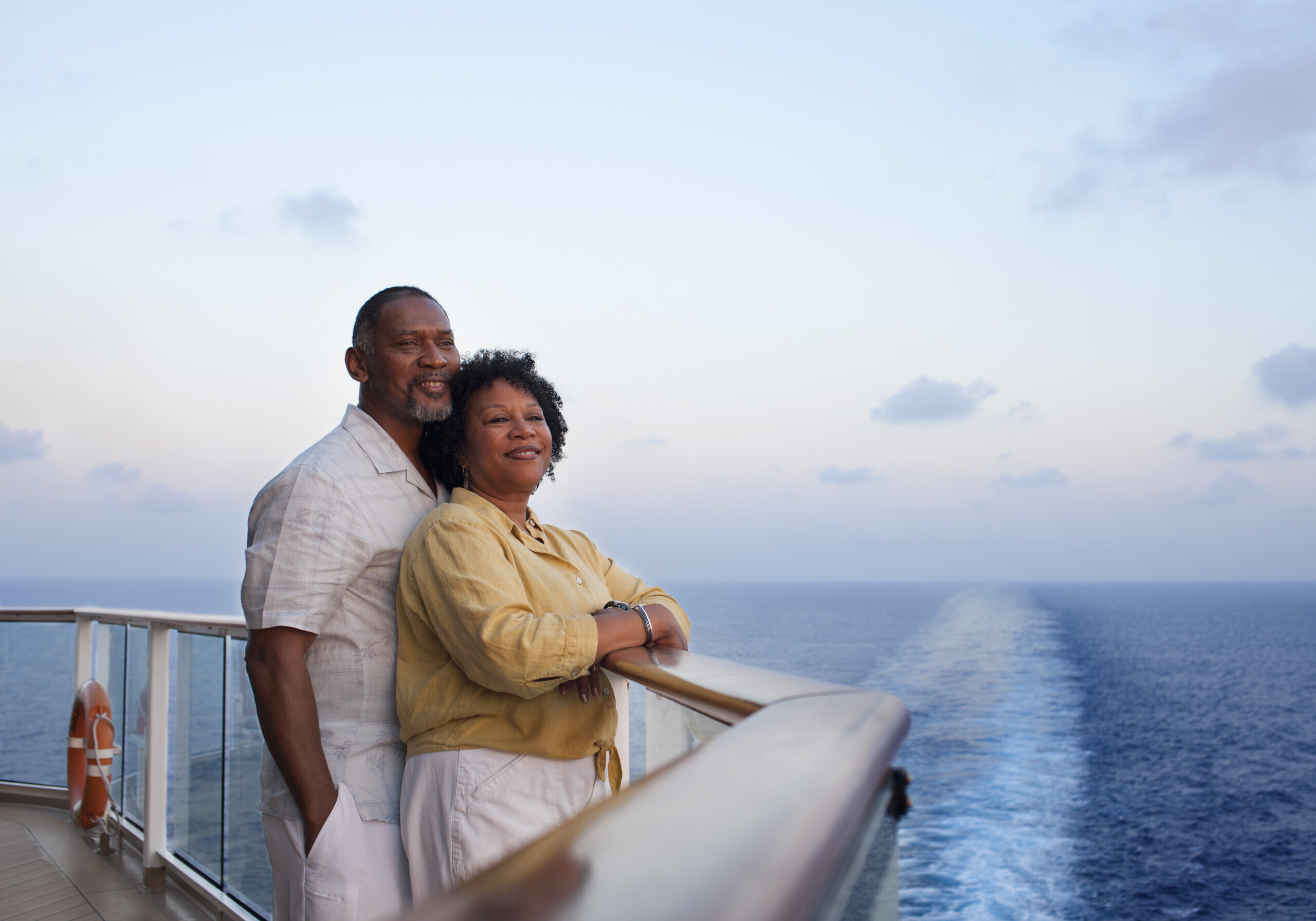 Mature African American couple, standing and leaning on railing on deck of a cruise ship, looking off into the distance.